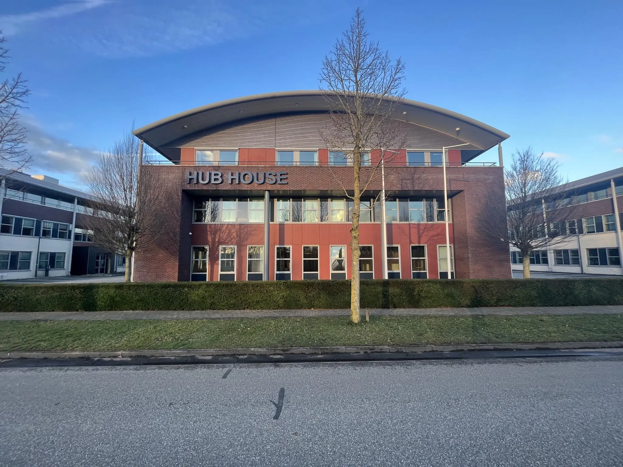 Vooraanzicht van het kantoorgebouw "HUB HOUSE" met rode en bakstenen gevel en kale bomen ervoor.